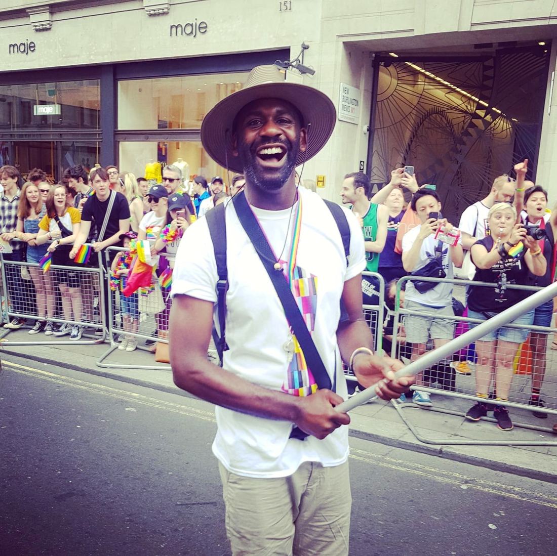 A man in a wide-brimmed hat laughing broadly at Pride, crowds and rainbow flags behind him